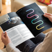 A person holding an open printed brochure n a bright indoor setting. The brochure has a black cover page with colorful abstract ribbon graphics and white interior pages with text and photos.