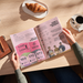 A person's hands holding an open magazine or book on a light wood table, brochure printed using 100% recycled paper stocks.