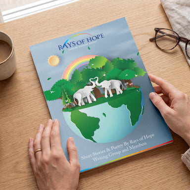 A person holds a printed erfect Bound Brochure titled "Rays of Hope Writing Workshop" on a wooden table. The cover shows a colorful illustration of elephants on a globe with trees, a rainbow, and a sun.