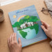 A person holds a printed erfect Bound Brochure titled "Rays of Hope Writing Workshop" on a wooden table. The cover shows a colorful illustration of elephants on a globe with trees, a rainbow, and a sun.