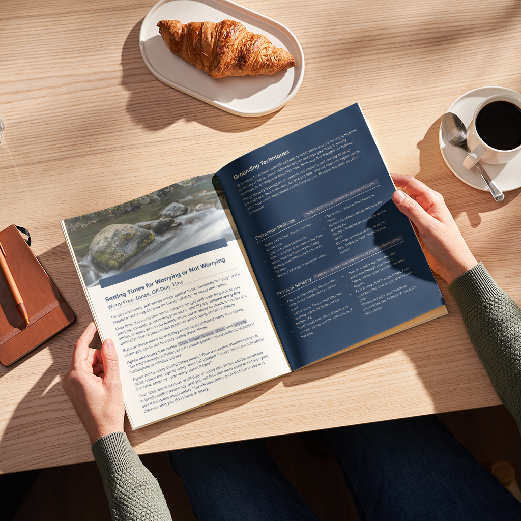 A person's hands holding an open book on a light wood table, with a croissant on a plate and a cup of coffee nearby in a warm, sunlit setting.